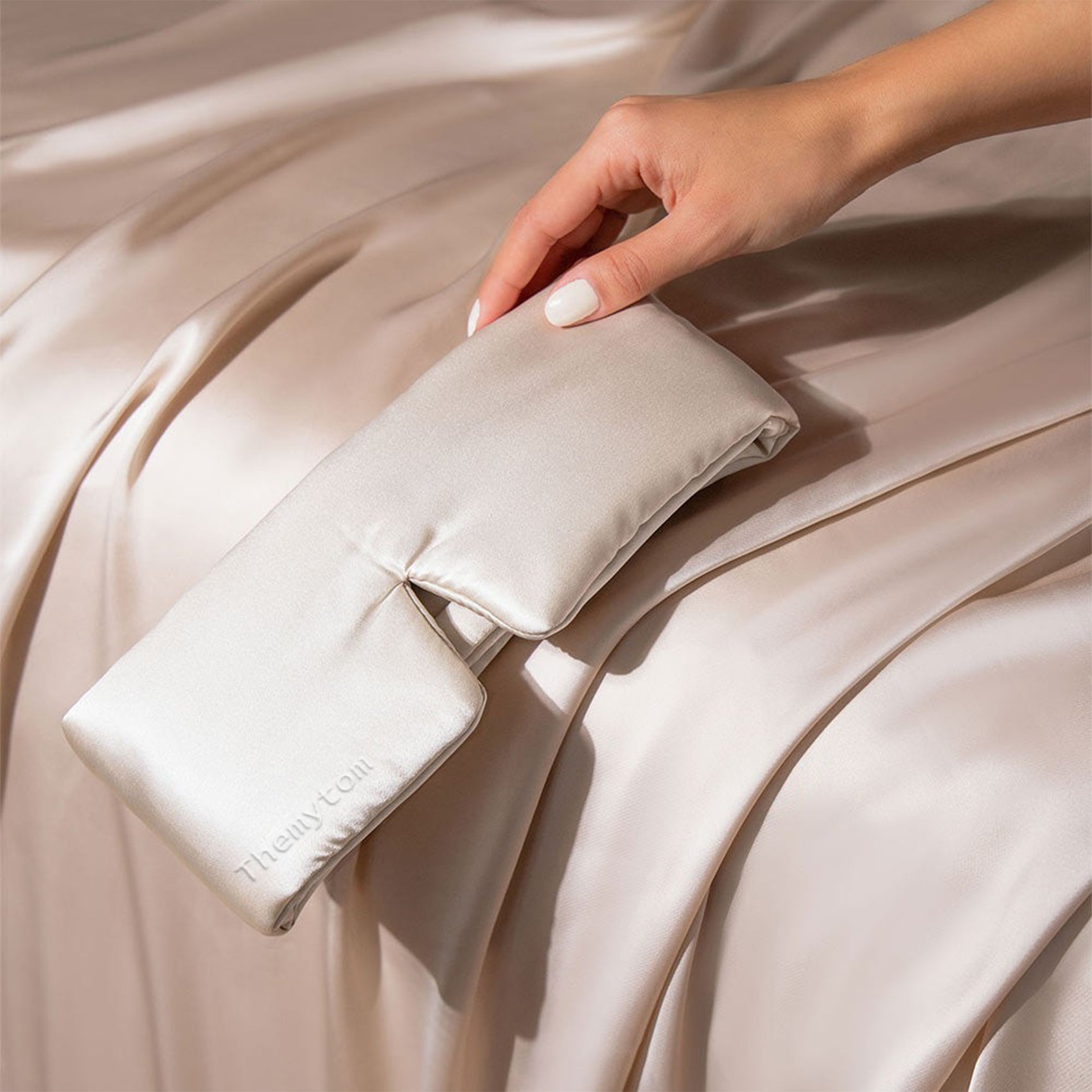 Folded beige satin pillowcase held by a hand on a beige fabric background Nº 08 Brume d’Argent